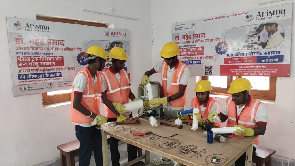 Four trainees in safety helmets and high-visibility vests handling tools and household equipment during an OES field technician vocational training session funded under CSR in India